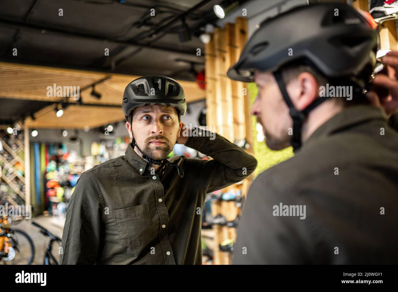 Il giovane uomo è venuto al negozio di biciclette. Sta misurando il casco. Il maschio sceglie il casco nel negozio di attrezzature sportive. Acquisto di nuovo Foto Stock