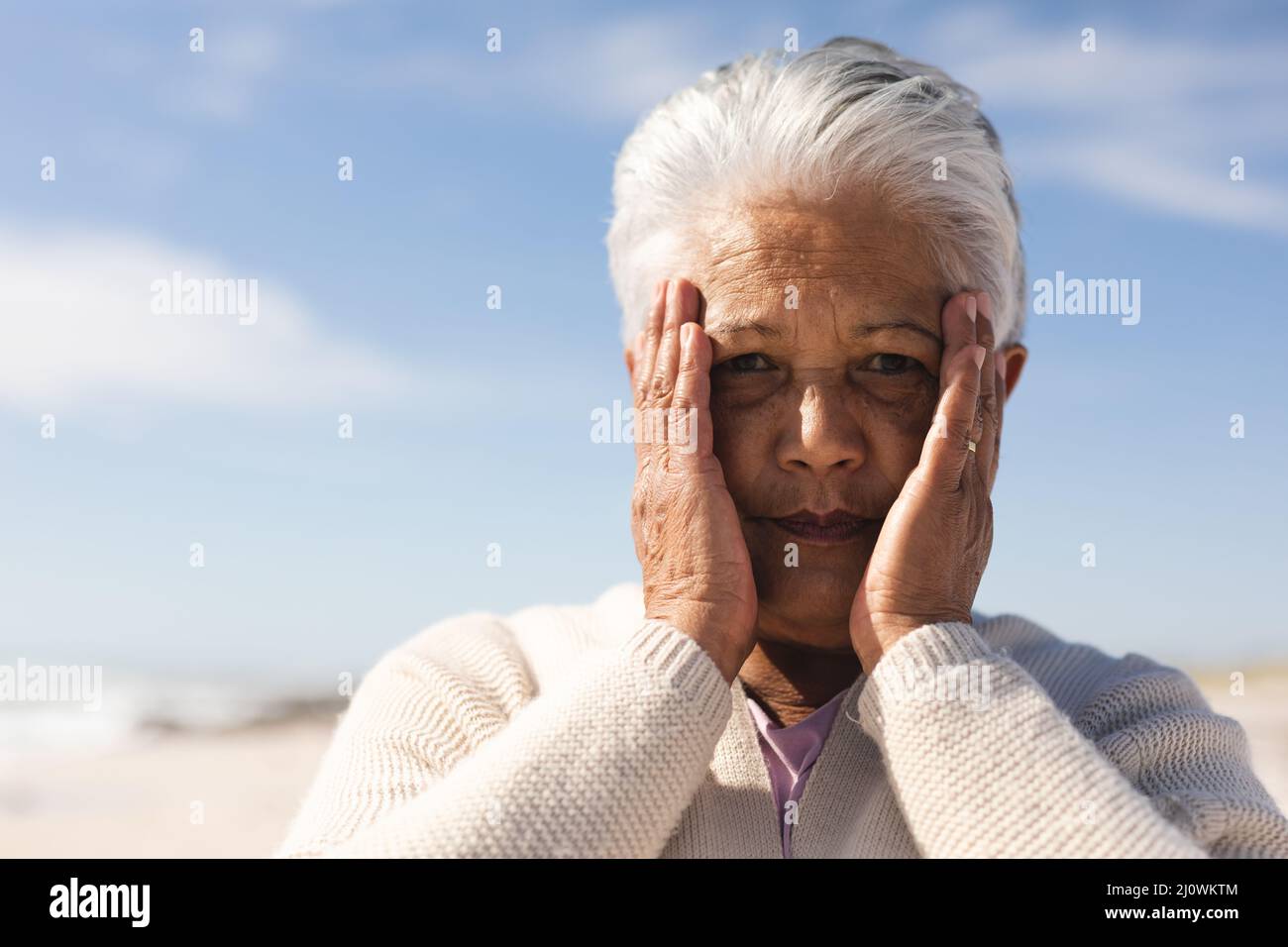 Ritratto di donna biraciale anziana preoccupata con le mani che ricoprono il viso in spiaggia in giorno di sole Foto Stock