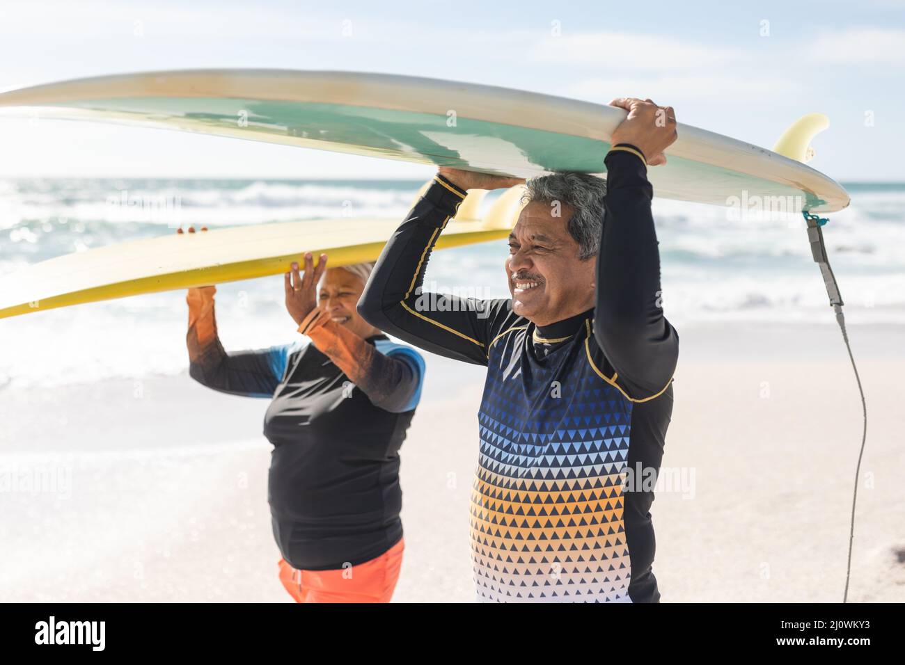 Coppia anziana multirazziale felice che trasporta tavole da surf sopra le teste alla spiaggia durante la giornata di sole Foto Stock