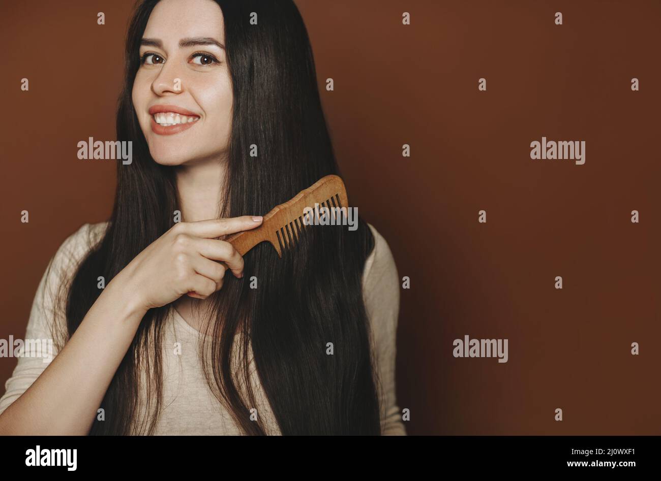 Giovane donna bruna felice pettinando i suoi capelli lunghi sani e sani e sorridendo alla macchina fotografica. Cura dei capelli Foto Stock