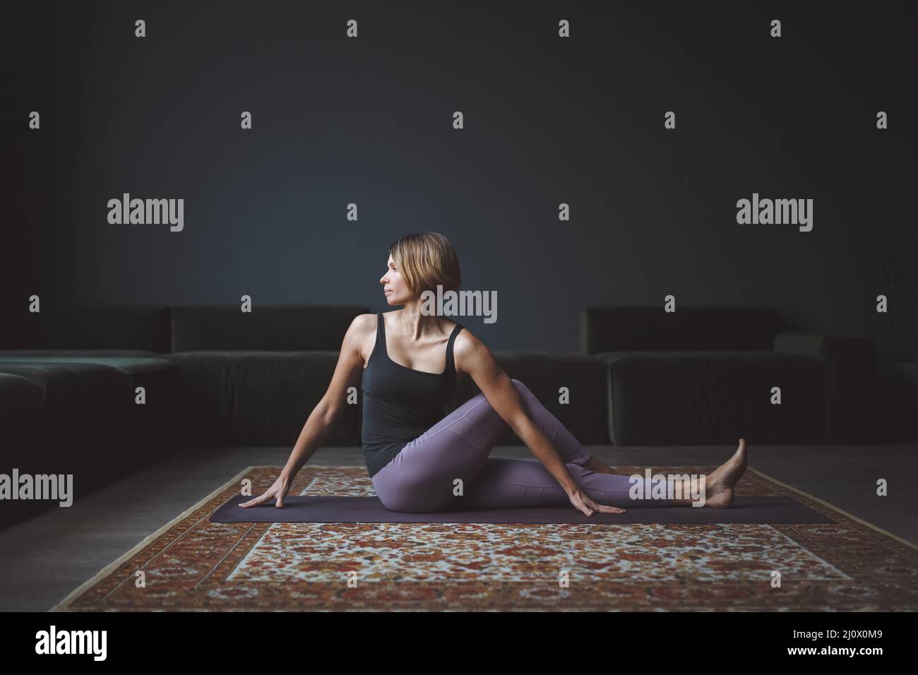 Giovane ragazza attraente che fa Ardha Matsyendrasana o apertura spinale twist yoga posa seduta sul tappeto a casa moderna interni Foto Stock
