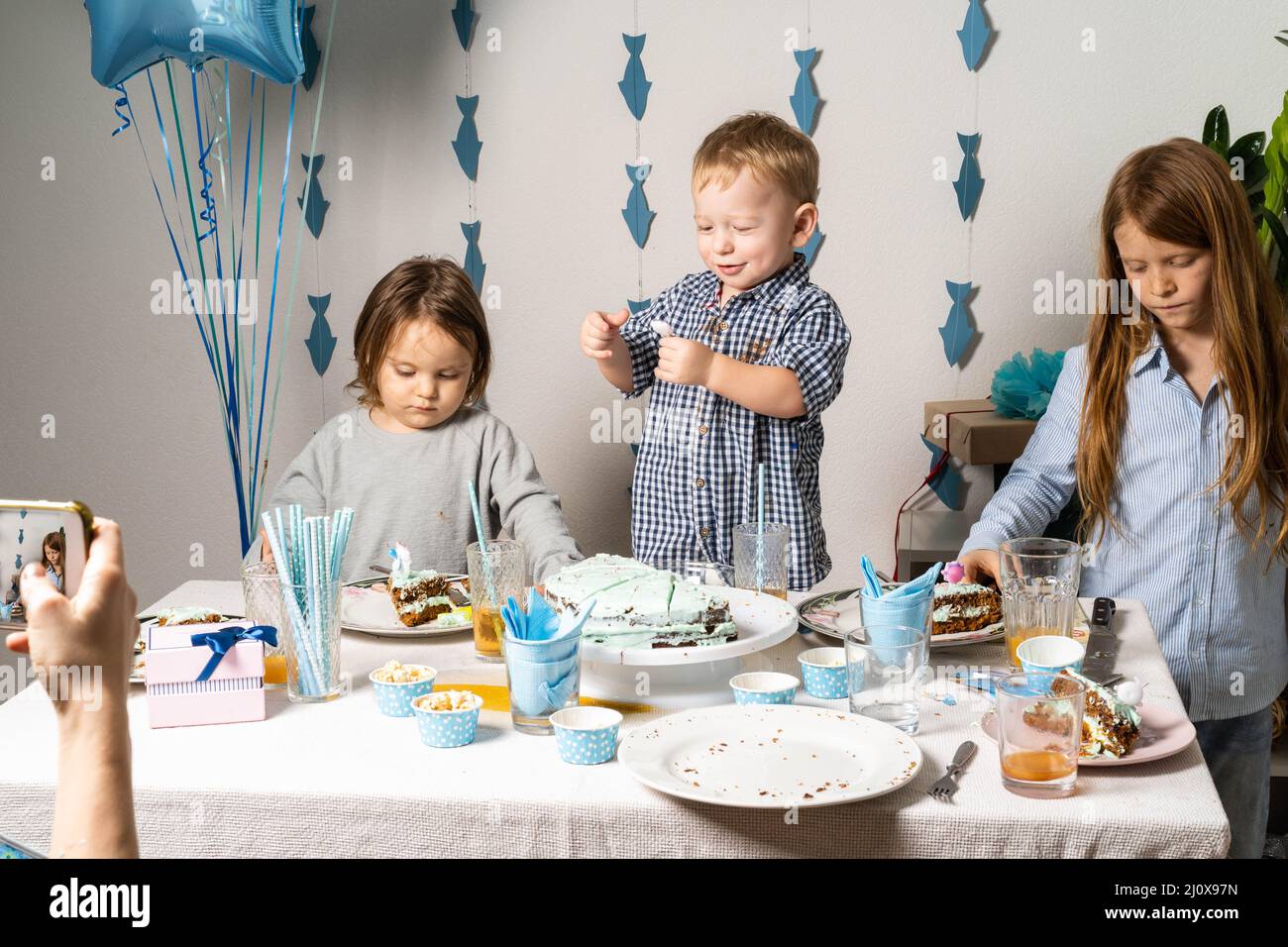 Fratelli. Compleanno del ragazzo al tavolo. I ragazzi al tavolo tagliano una torta di compleanno. Mamma scatta foto del compleanno del bambino al telefono. Foto Stock