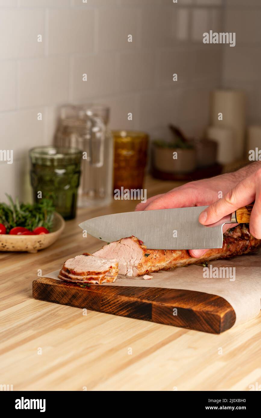 Taglio uomo irriconoscibile con filetto di maiale cotto a coltello grande sul tagliere, verticale Foto Stock