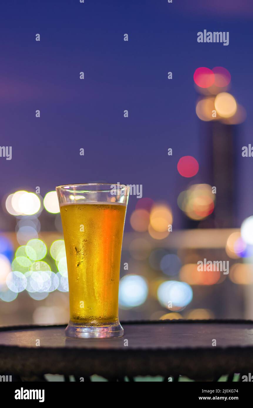 Un bicchiere di birra mette sul tavolo al bar sul tetto con sfondo luminoso bokeh colorato della città. Foto Stock