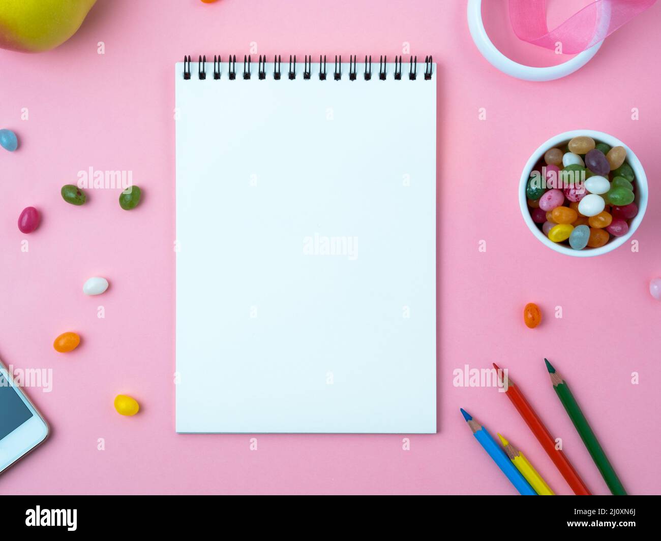 Notebook aperto con un foglio bianco pulito, dolci, telefono cellulare, pastello, decorazioni su un tavolo rosa luminoso Foto Stock