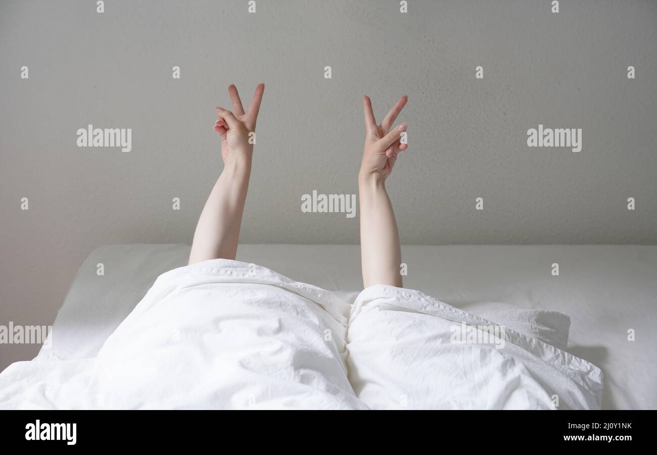 donna che mostra la pace o v mano segno mentre sdraiato in letto accogliente e nascosto sotto le coperture Foto Stock