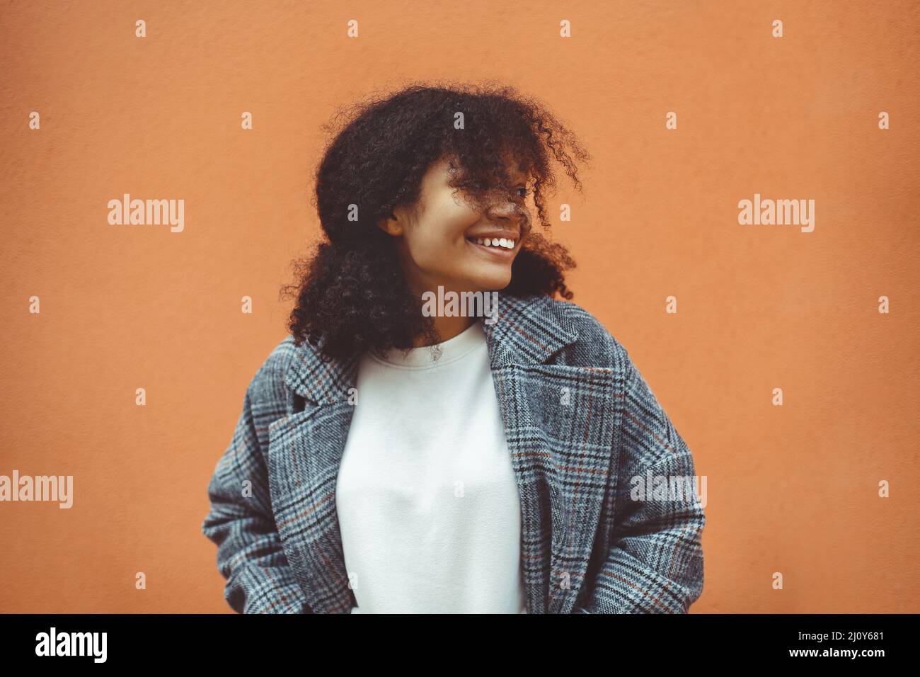 Felice etnia africana ragazza con capelli ricci scuri in elegante cappotto guardando da parte e sorridente Foto Stock
