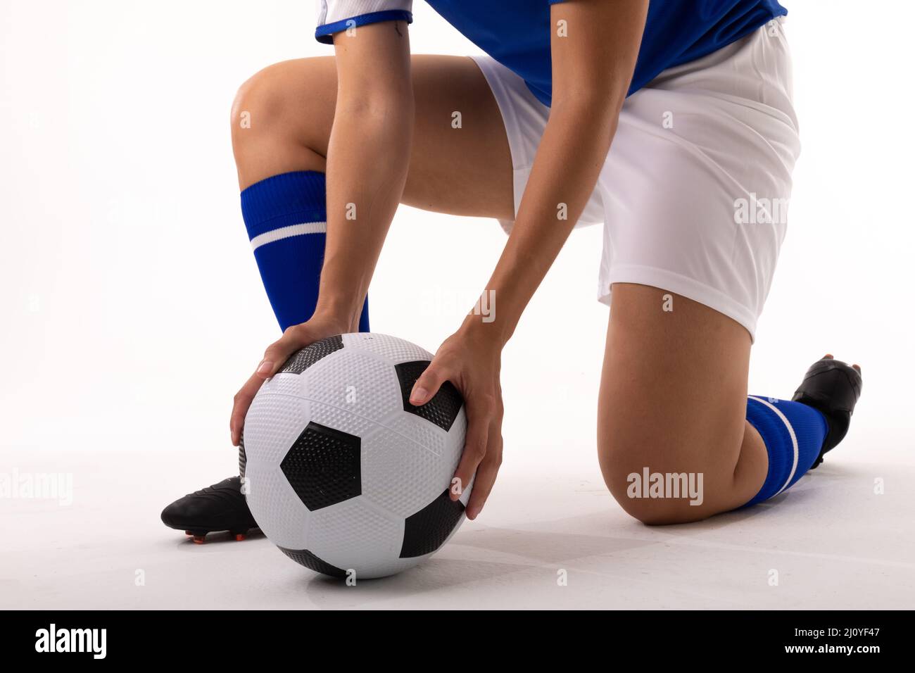 Sezione bassa di giovane donna biraciale che tiene la palla di calcio mentre si inginocchiano su sfondo bianco Foto Stock