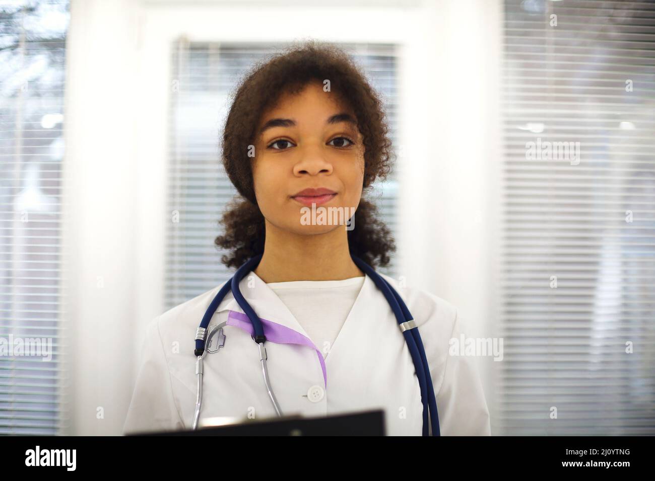 Felice medico africano americano medico donna indossare uniforme medica con stetoscopio intorno al collo sorridendo alla macchina fotografica durante il turno di lavoro in clin moderno Foto Stock