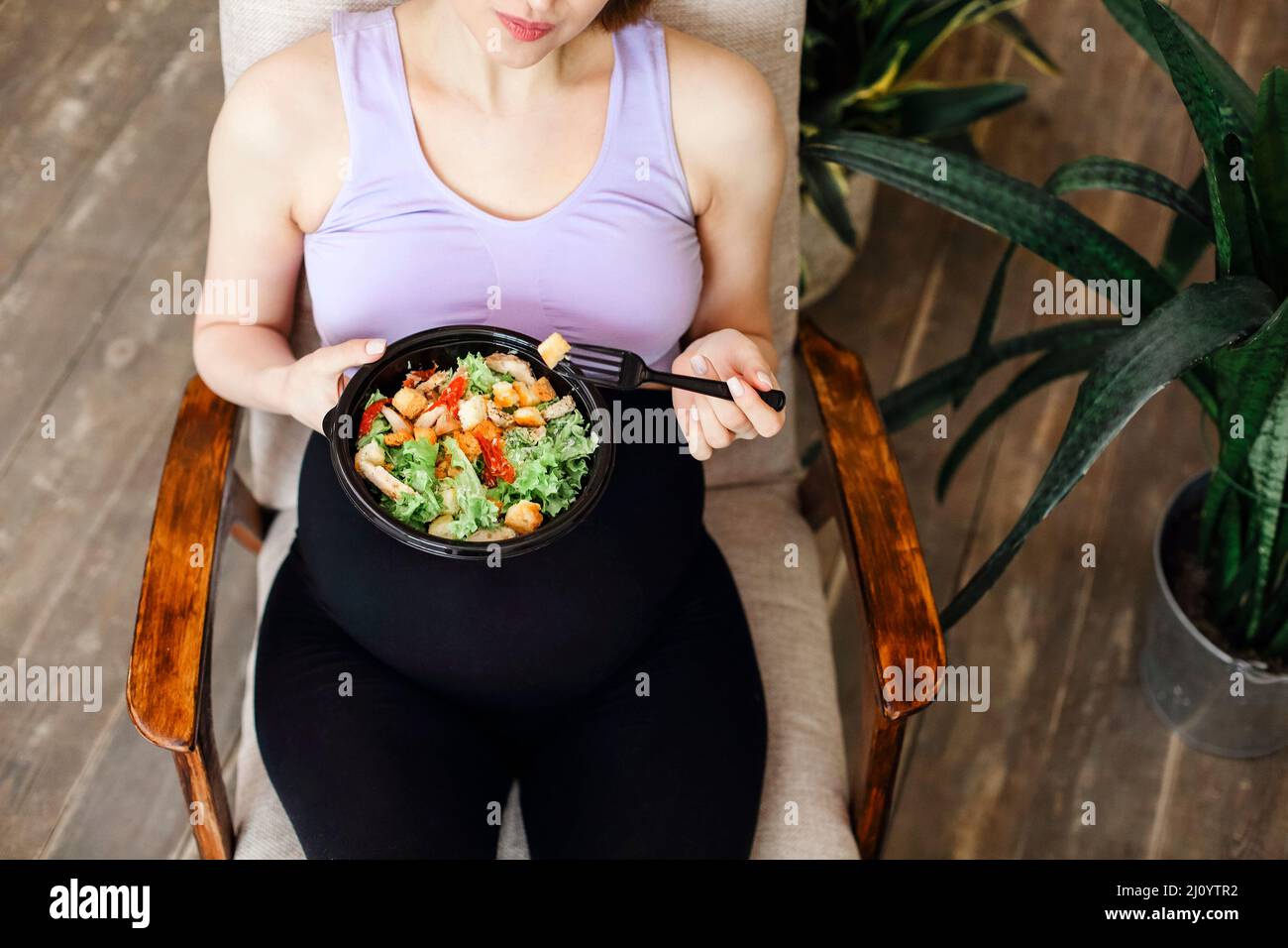 Donna bruna giovane incinta che mangia insalata sana dalla vista dall'alto della ciotola. Cibo sano mentre si pranzano su una comoda poltrona Foto Stock