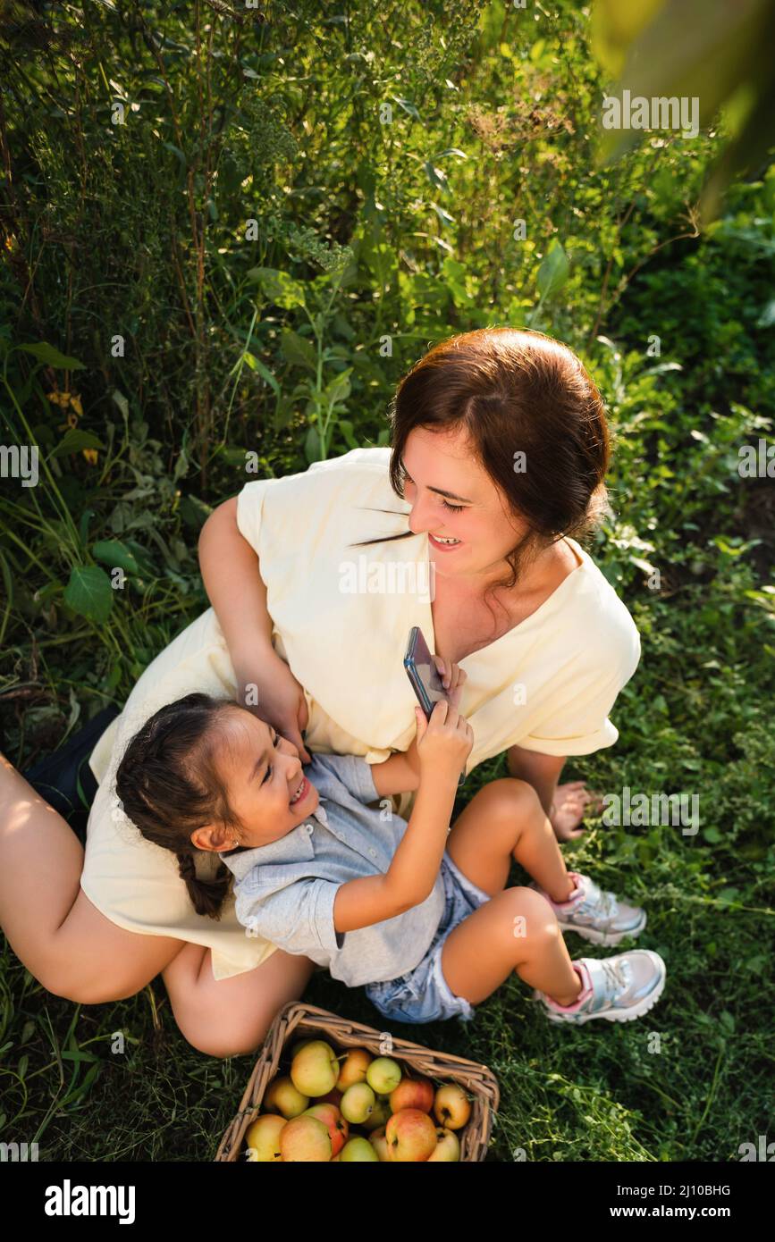 La madre caucasica e la figlia asiatica prendono un selfie nel frutteto in estate Foto Stock