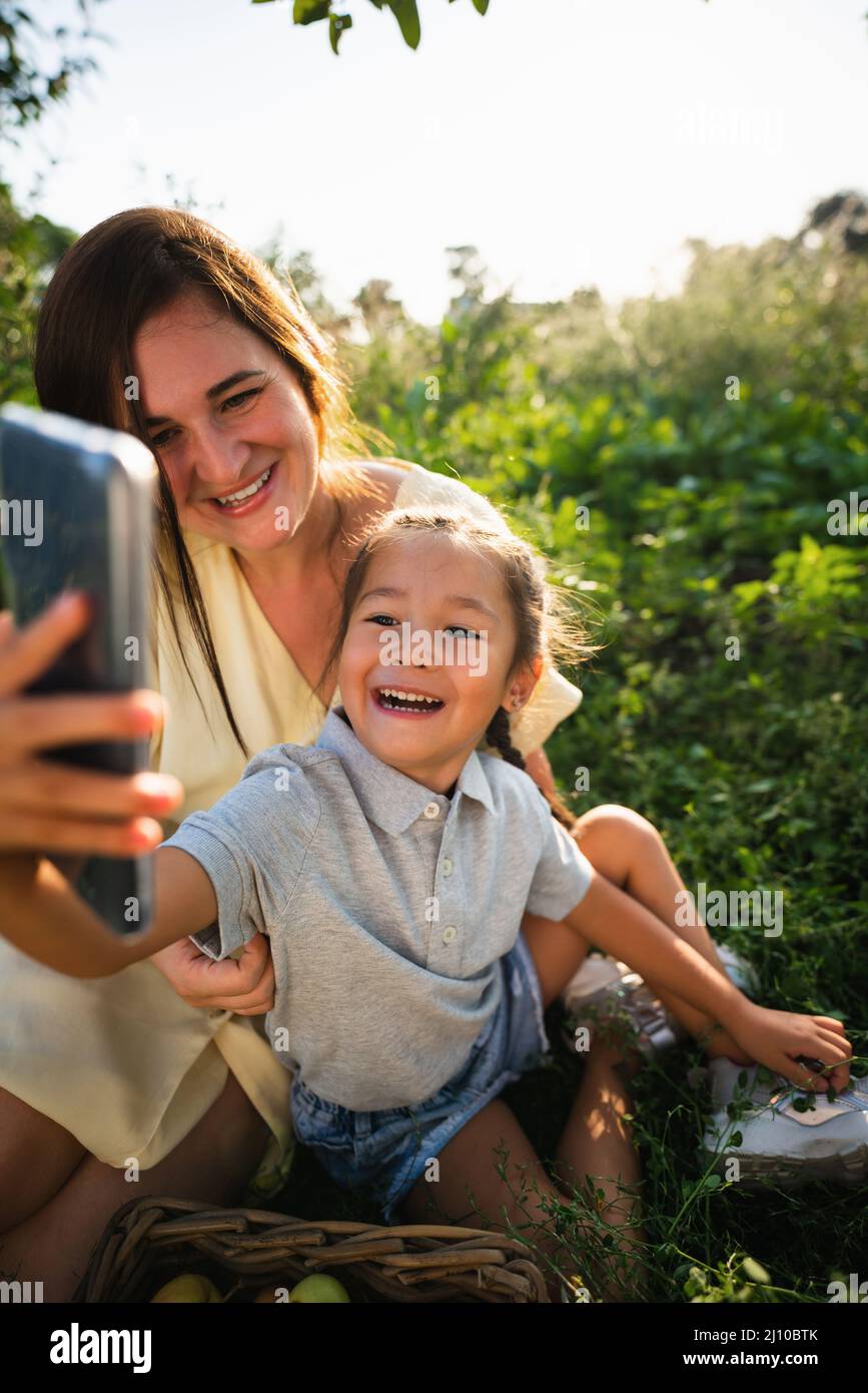 La madre caucasica e la figlia asiatica prendono un selfie nel frutteto in estate Foto Stock