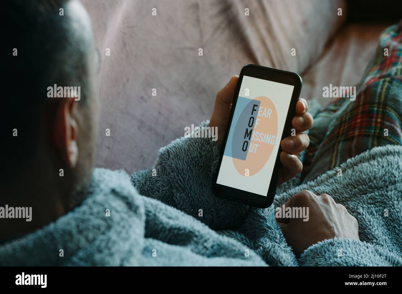 un uomo caucasico, sdraiato sul divano con pigiami e vestito di casa, guarda al suo smartphone, che legge il testo fomo, per paura di perdere fuori Foto Stock