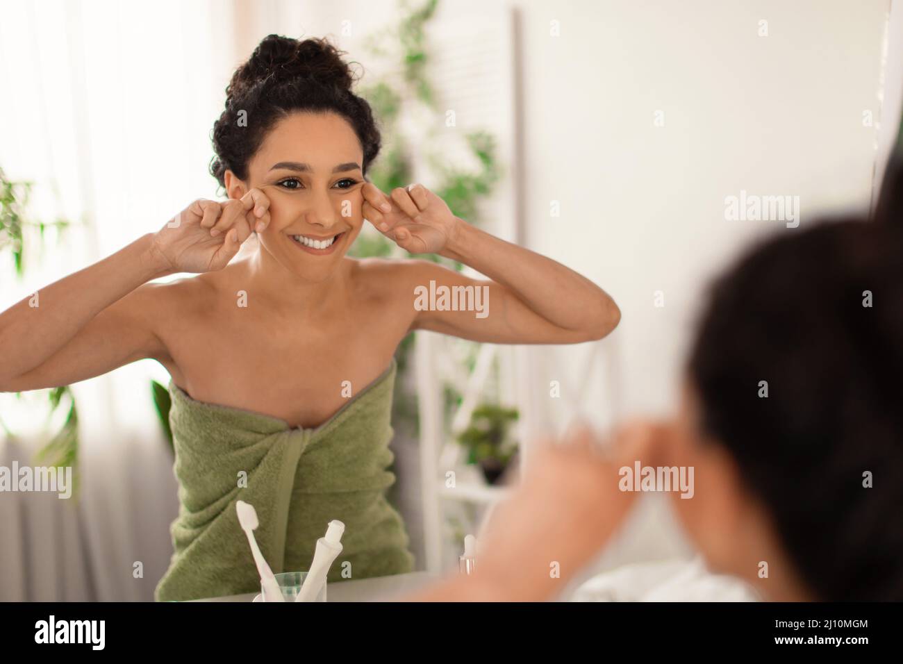 Bella giovane donna che fa linfodrenaggio viso auto massaggio vicino specchio a casa, spazio libero Foto Stock