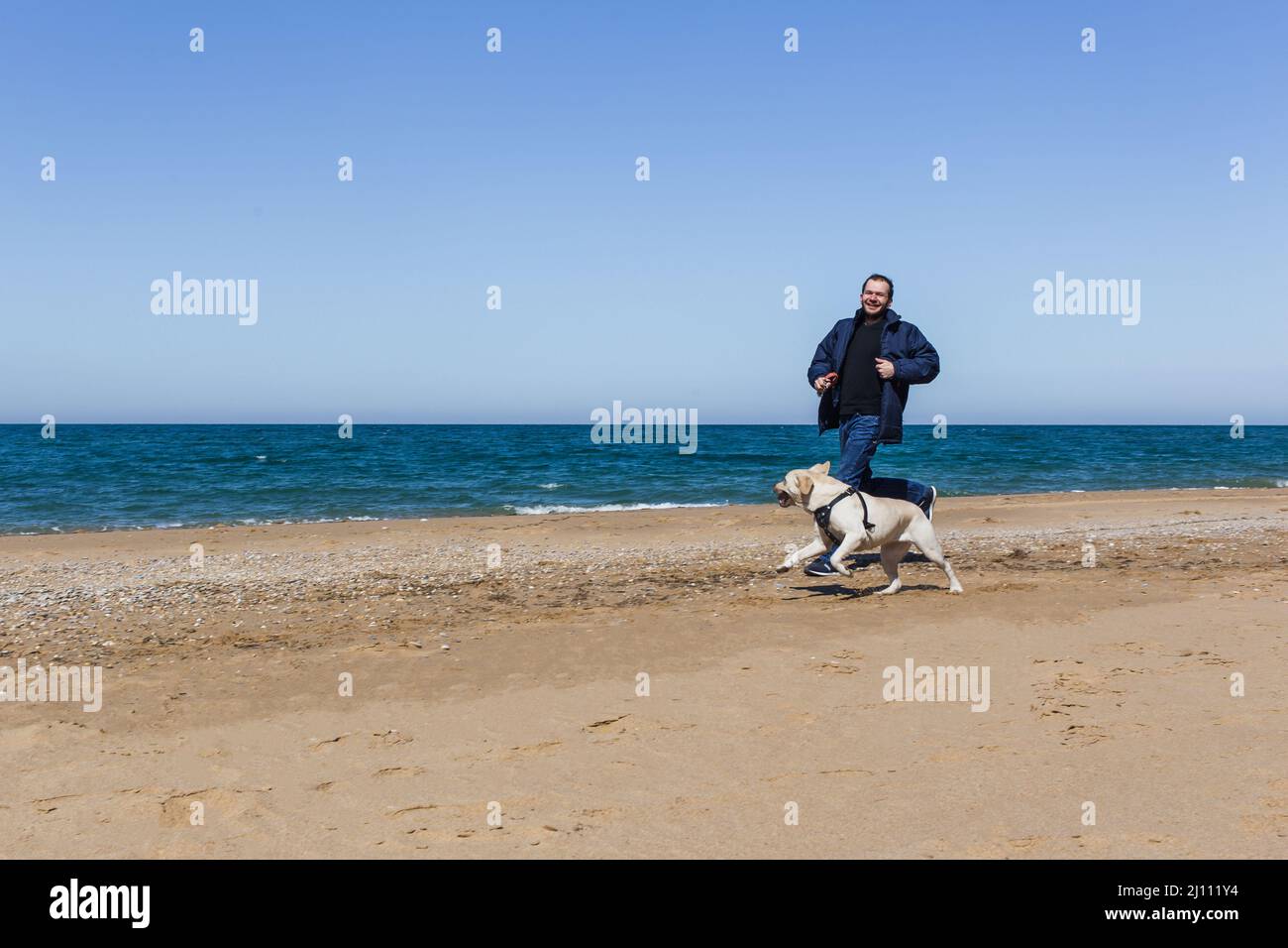 Un uomo in inverno vestiti corre lungo una spiaggia di sabbia con un cane in munizioni Foto Stock