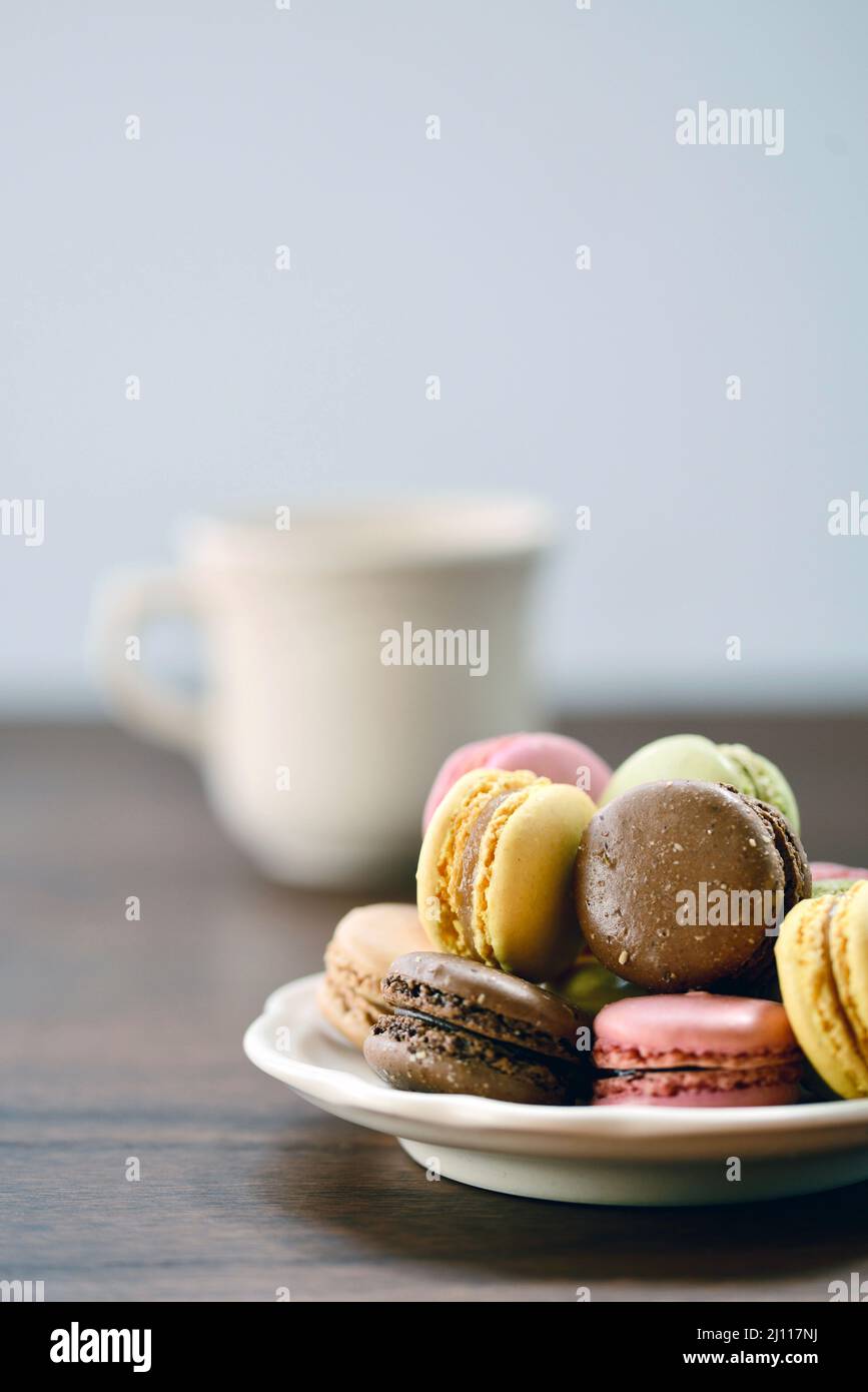 Un mucchio di biscotti francesi di macaron accatastati su un piatto su un tavolo di legno in luce naturale con una tazza di latte o caffè. Foto Stock Un mucchio di biscotti francesi di macaron accatastati su un piatto su un tavolo di legno in luce naturale con una tazza di latte o caffè. Foto Stock