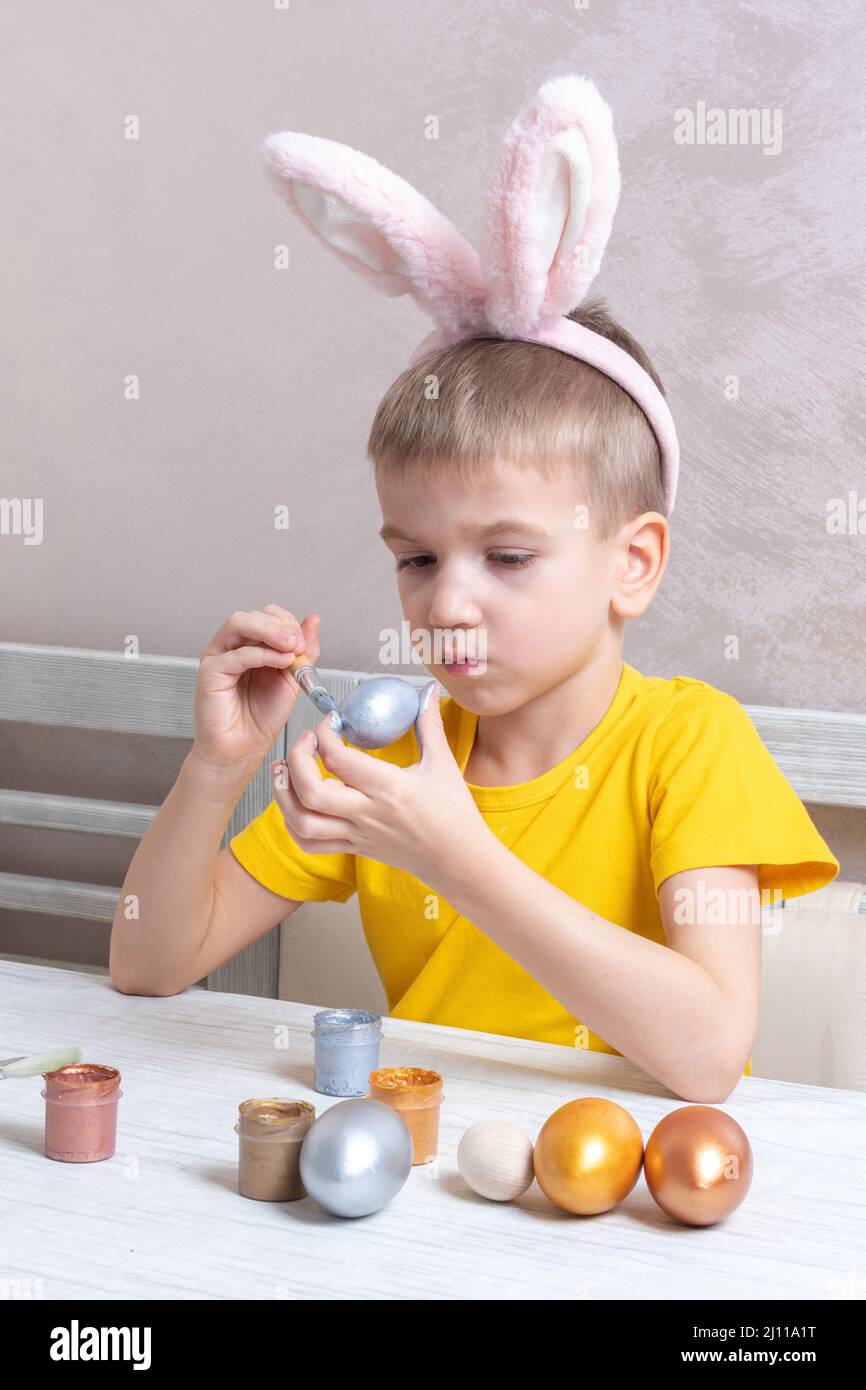 Un ragazzo biondo in coniglietto orecchie dipinge le uova per le vacanze di Pasqua nella cucina di casa, cornice verticale. Il bambino si diverte e festeggia la vacanza. Foto Stock