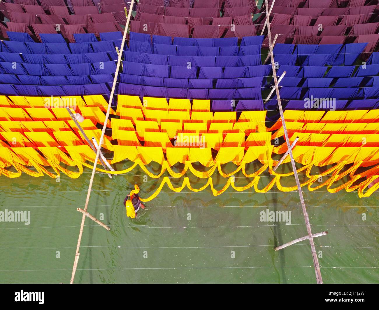 I lavoratori appendono migliaia di tessuti colorati su fili di ferro legati tra un telaio di bambù e costantemente li trasformano in modo che si asciughino perfettamente in un campo allagato a Narayanganj, Bangladesh. Questa è la parte finale del processo di tintura dopo che il panno è fatto in t-shirt e giubbotti presso la fabbrica di abbigliamento. I panni vengono tinti utilizzando macchine in una fabbrica e disposti in campi vicini in quanto è più economico e sostenibile asciugarli alla luce del sole. Dopo essere stati lasciati al sole fino a 5-6 ore, vengono poi fatti in magliette, giubbotti e abiti. Bangladesh. Foto Stock