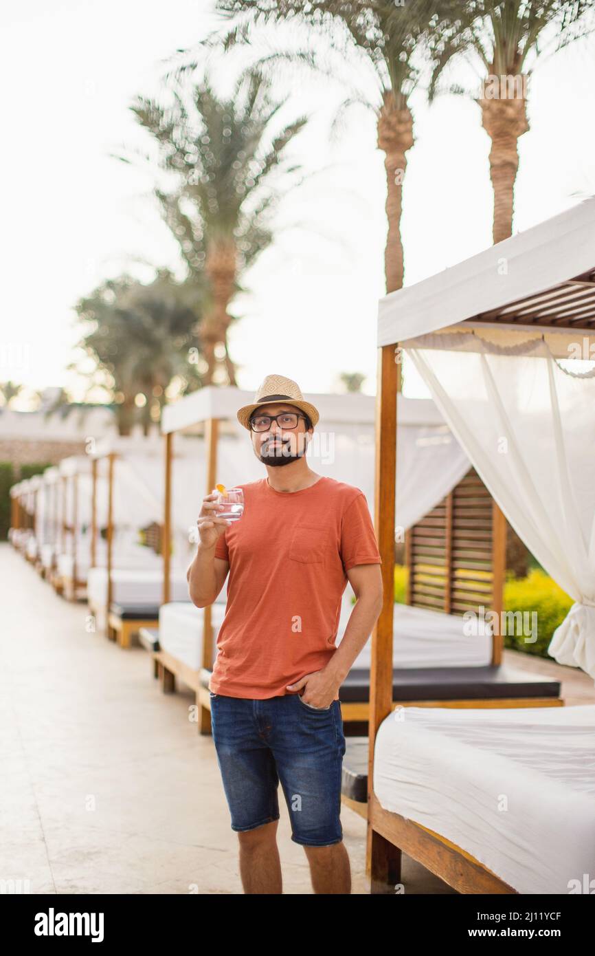 Un giovane uomo bello in una T-shirt arancione, cappello di paglia e  pantaloncini in denim stand, bere fresco limonata, guardando via sullo  sfondo di gazebo in un resort tropicale di lusso Foto