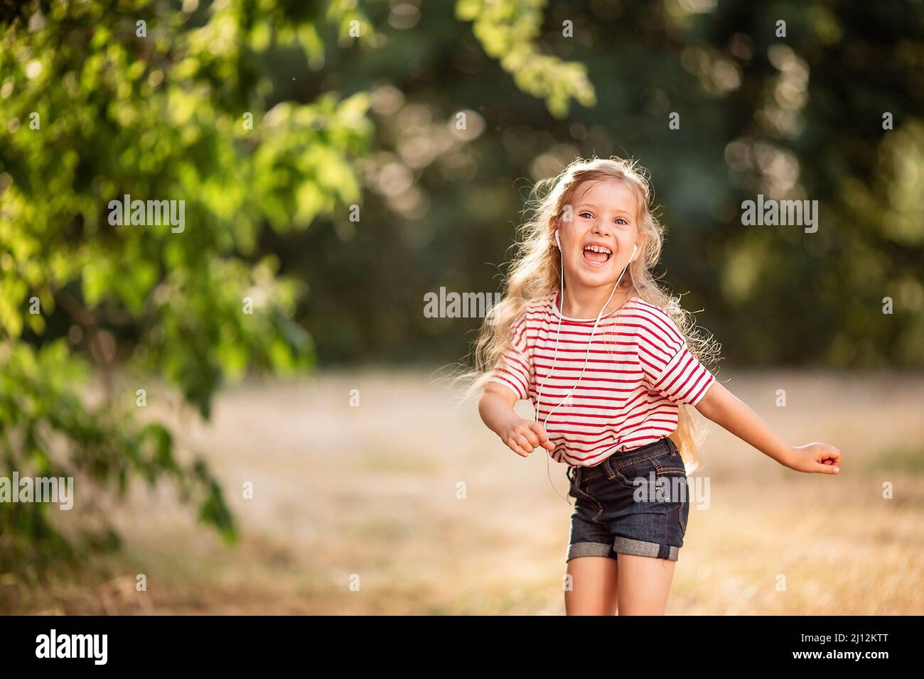 Buona ragazza bionda che ascolta musica con le cuffie, ballando e cantando in natura nel Parco. Foto Stock