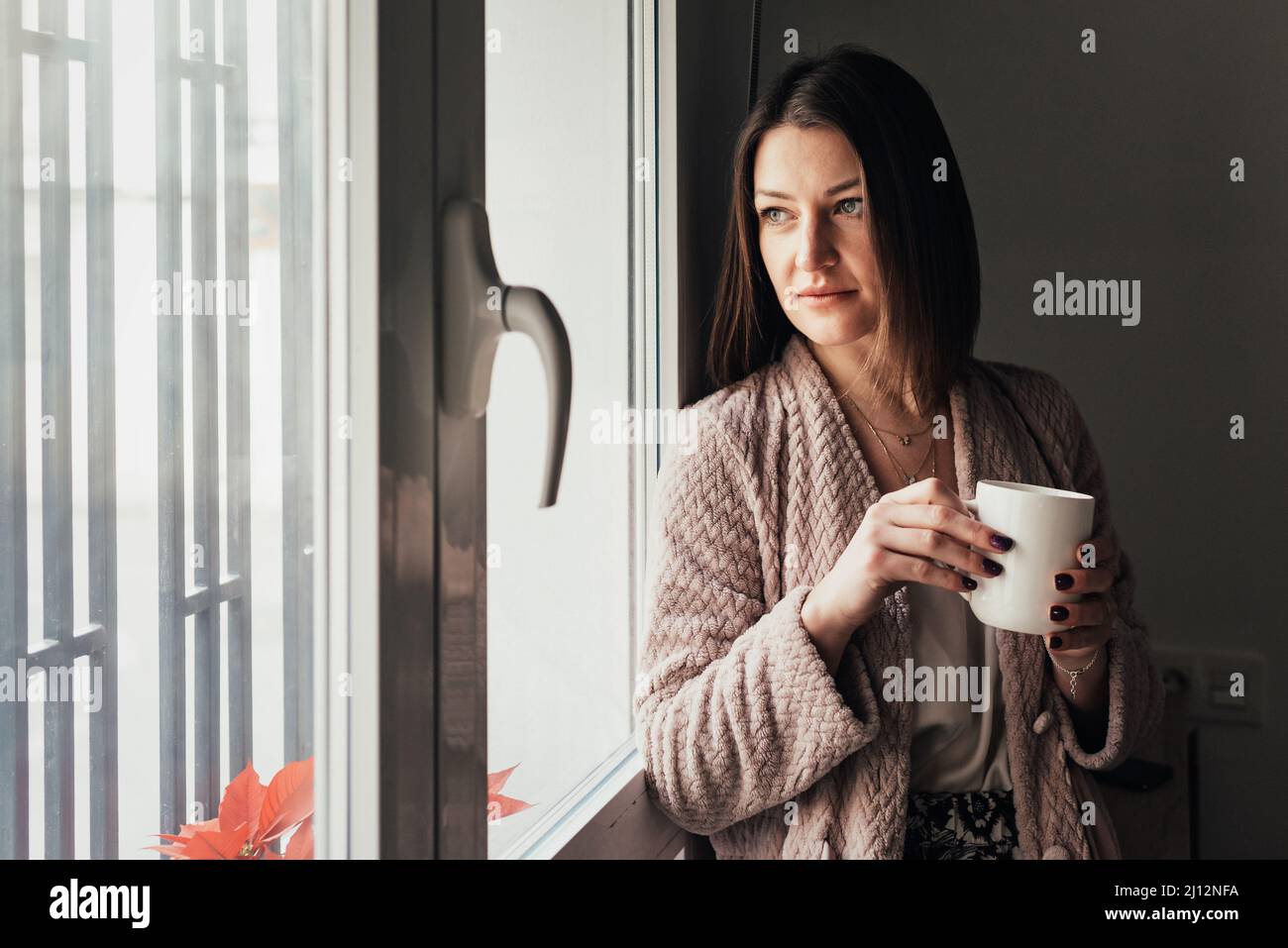 Donna rilassata che tiene una tazza di caffè mentre guarda attraverso una finestra. Foto Stock
