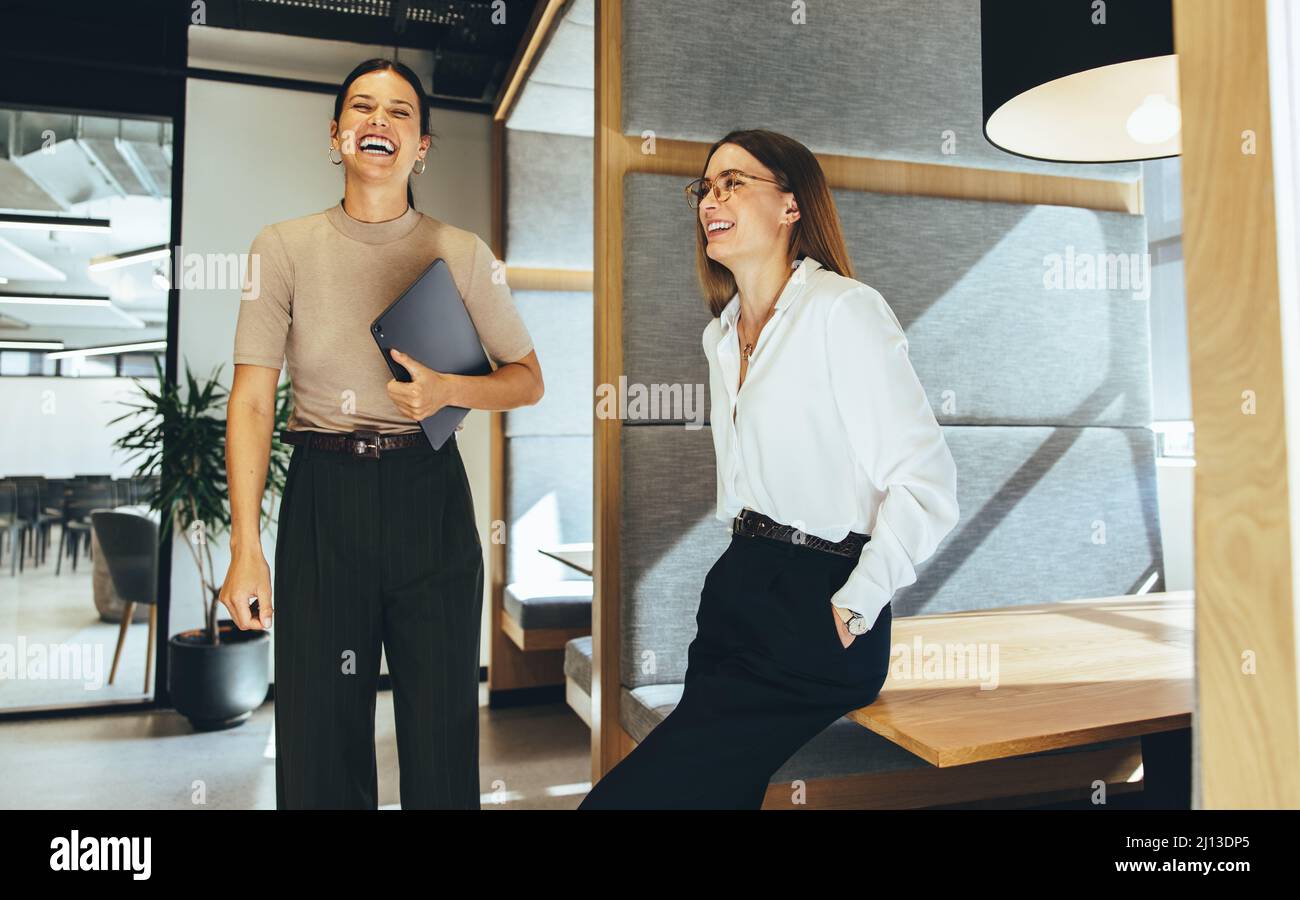 Professionisti di affari femminili che ridono allegro in un ufficio. Due giovani donne d'affari di successo che fanno una breve pausa mentre insieme in un wor moderno Foto Stock