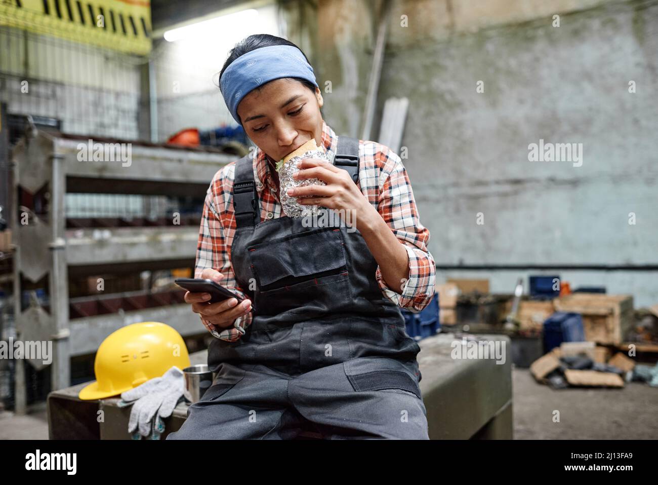 Giovane ingegnere ispanico ispanico affamato in tute e cerchietto blu che mordicchiava appetitosi sandwich durante il pranzo a pausa Foto Stock