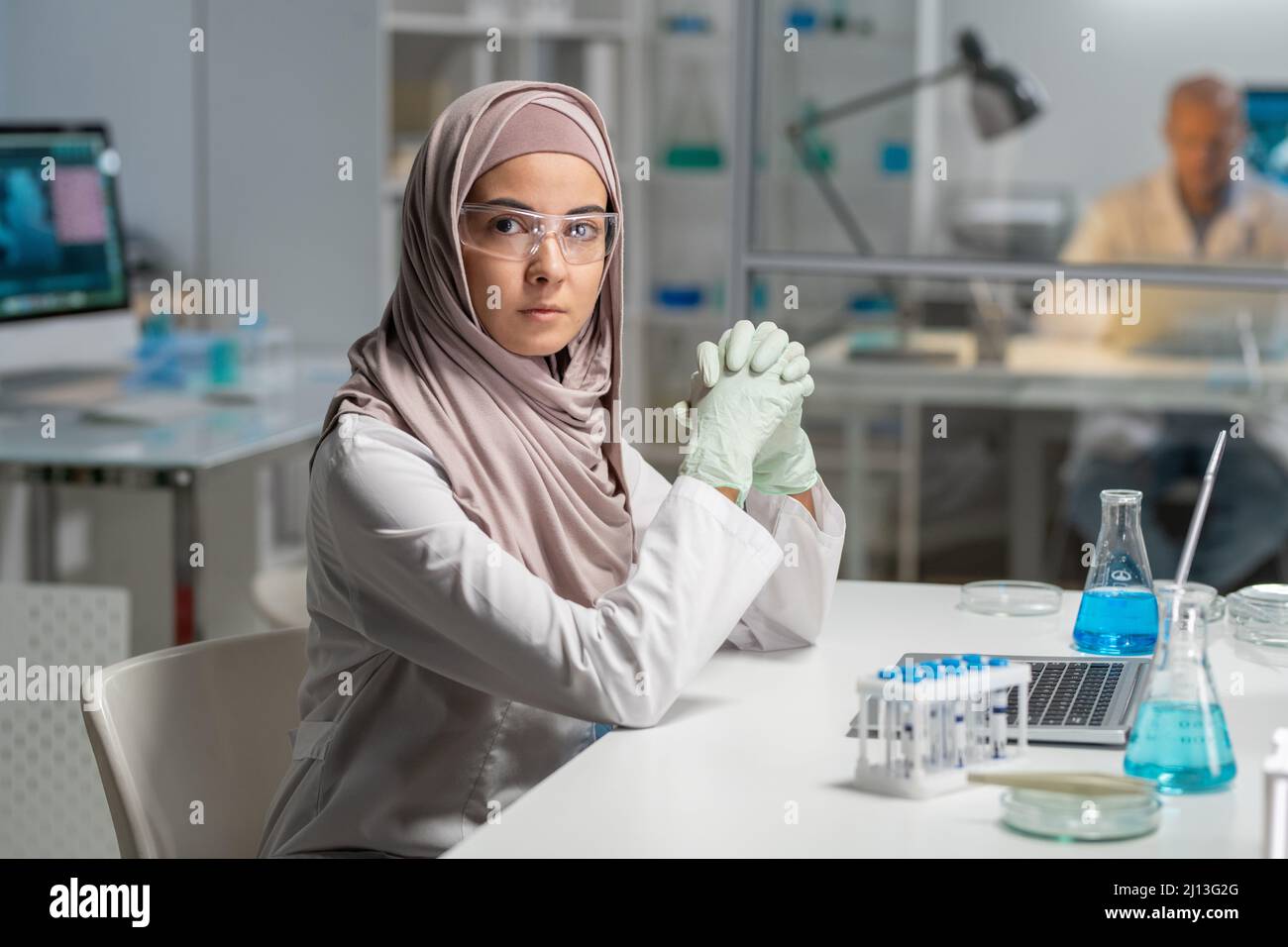 Giovane ricercatrice femminile contemporanea in occhiali protettivi e hijab guardando la macchina fotografica mentre si siede dalla scrivania di fronte al computer portatile Foto Stock