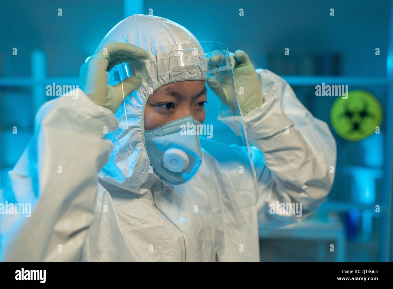 Giovane scienziata asiatica in tuta a rischio biologico, guanti e respiratore mettendo schermo protettivo sul viso di fronte al laboratorio Foto Stock