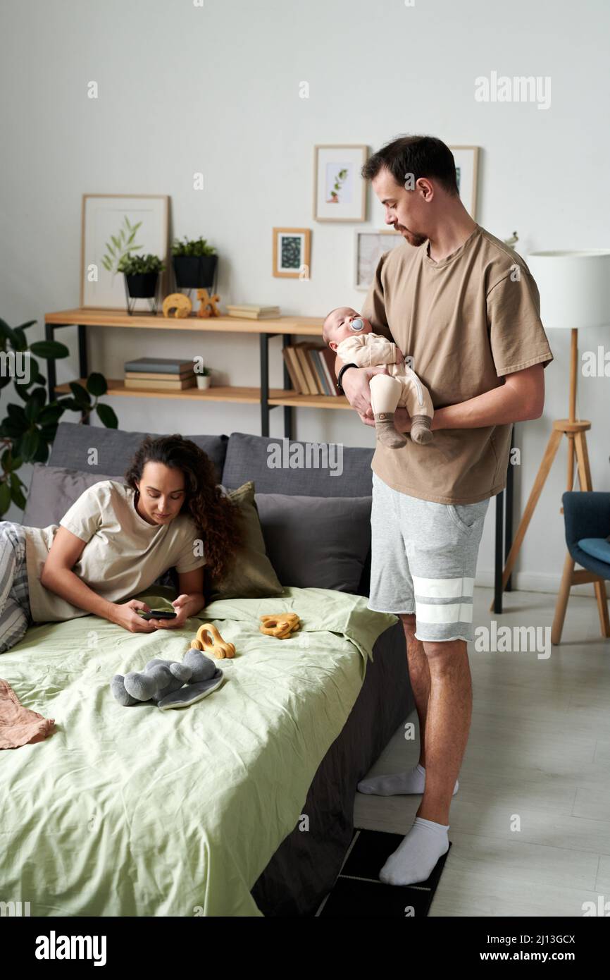 Giovane uomo in casualwear che tiene il bambino a dormire sulle mani mentre sua moglie riposa sul letto doppio e texting in smartphone Foto Stock