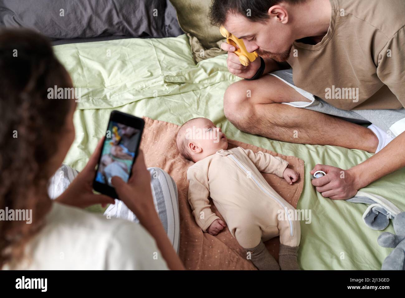 Giovane donna con smartphone fotografando il marito che gioca con il suo adorabile figlio sul letto matrimoniale a piacere Foto Stock