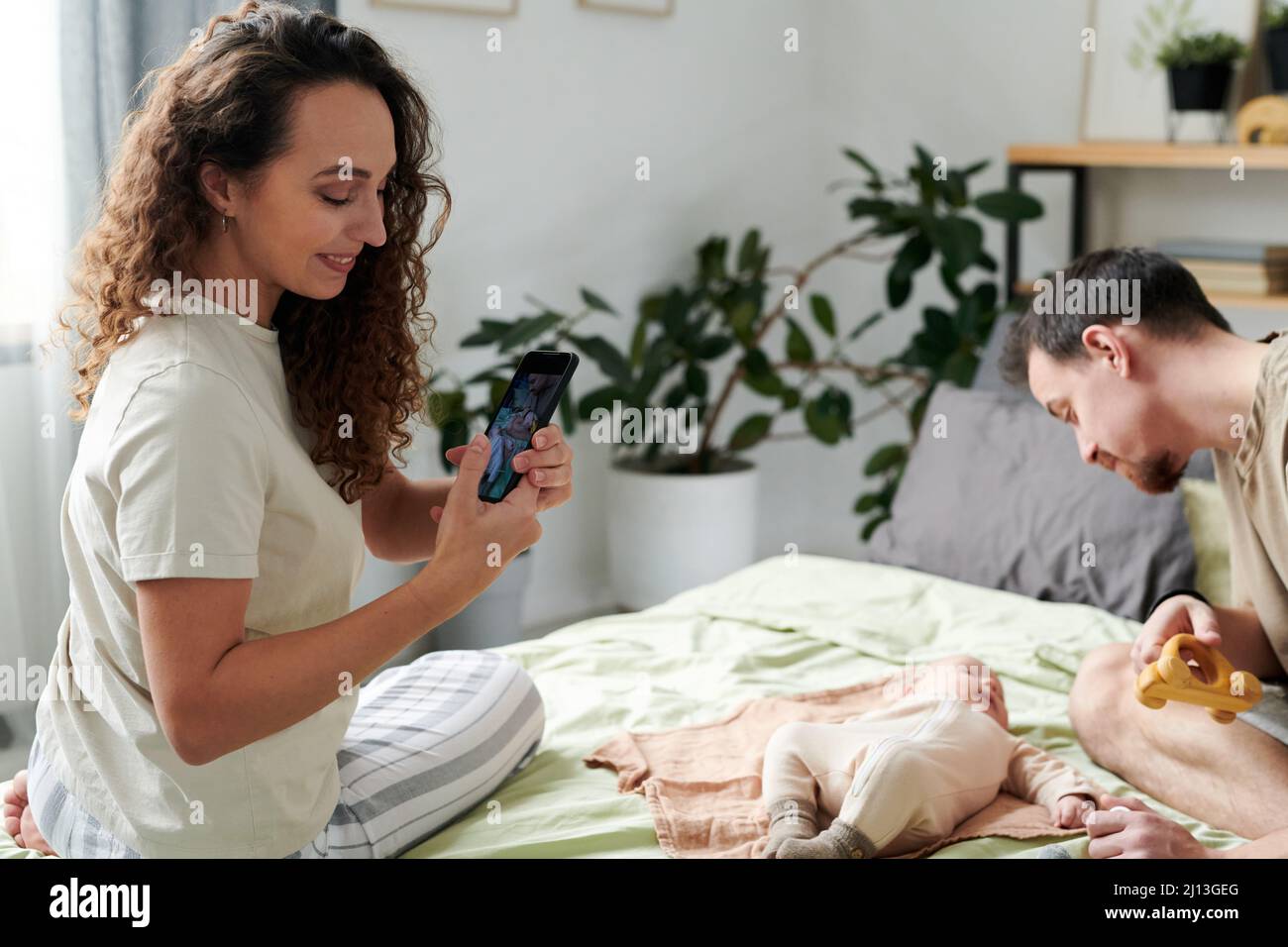 Giovane donna sorridente con telefono cellulare seduto sul letto matrimoniale di fronte al marito e il loro bambino simpatico figlio mentre si scatta la foto Foto Stock
