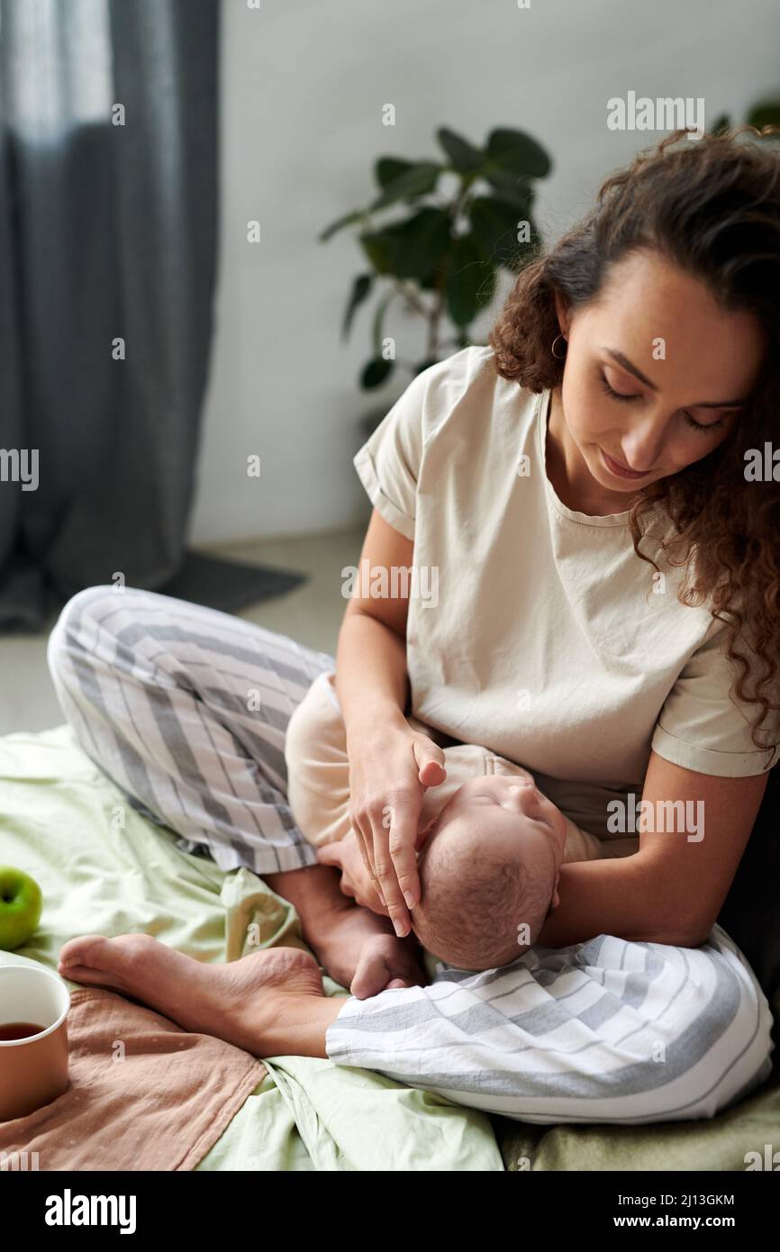 Giovane donna bruna che guarda il bambino che dorme sulle sue mani mentre si siede sul letto matrimoniale la mattina e facendo colazione Foto Stock