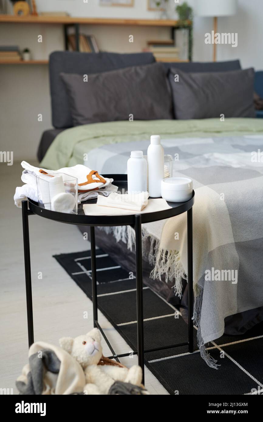 Set di bottiglie in plastica bianca con lozioni per il corpo e creme, pannolini e tamponi in cotone in contenitore in vetro su tavolino contro letto doppio Foto Stock