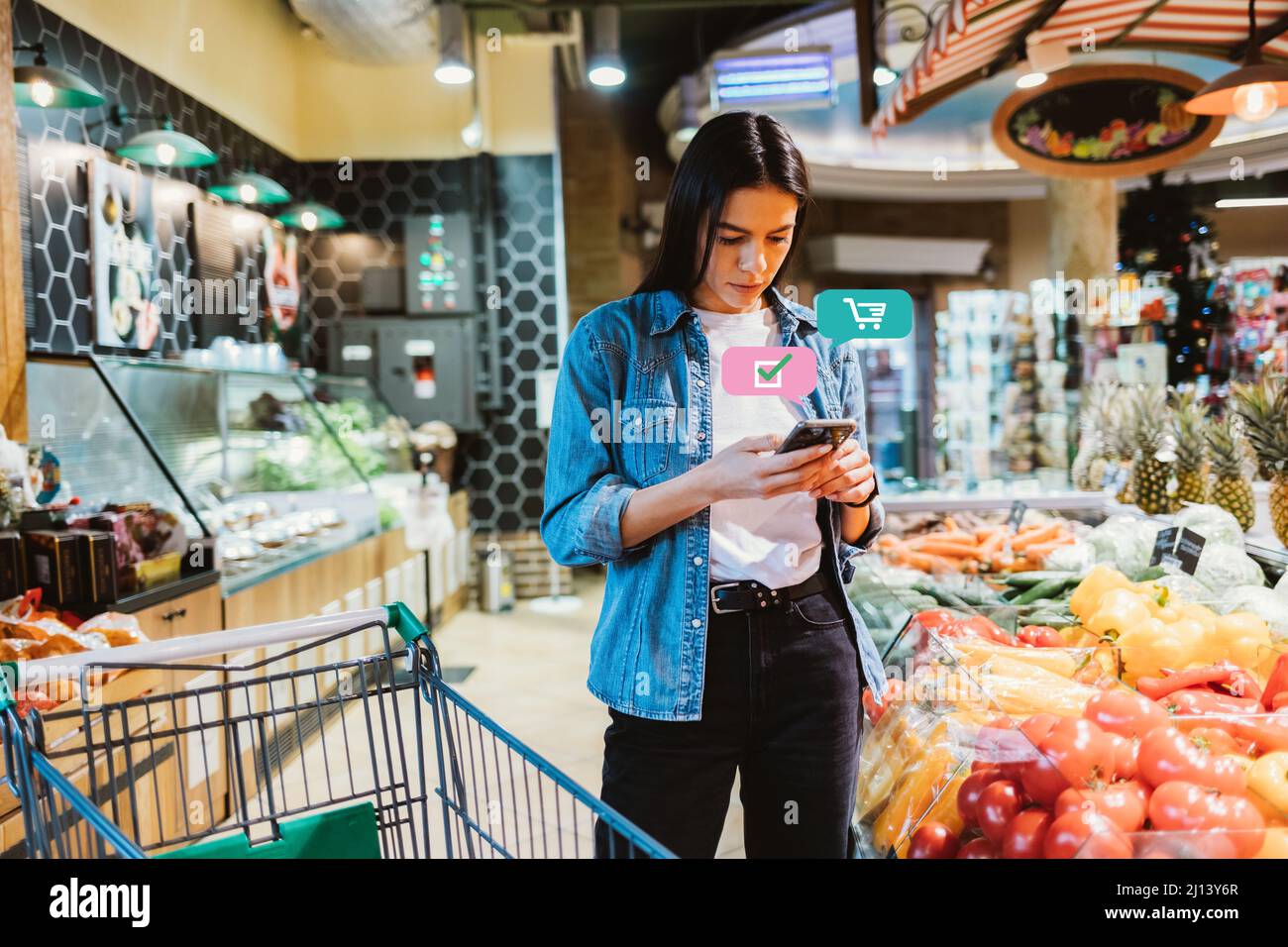 Shopping list app concept, giovane donna in supermercato guarda il suo smartphone per marcare i prodotti di cui ha bisogno Foto Stock