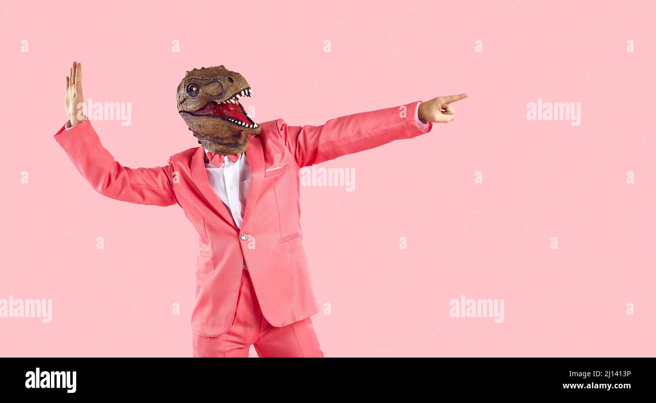 Pazzo ragazzo in abito da festa e maschera di dinosauro danzare e puntare via isolato su sfondo rosa Foto Stock
