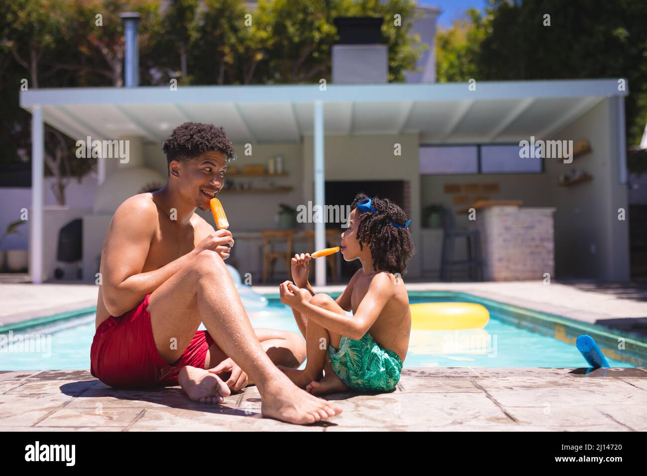 L'intera lunghezza del padre e del figlio ispanici che mangiano schiocchi di ghiaccio mentre si siedono insieme a bordo piscina Foto Stock