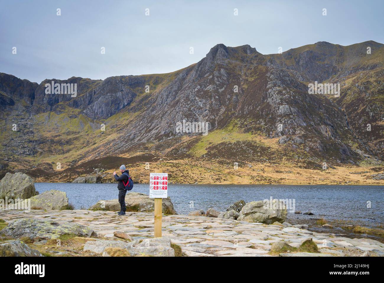 Vista laterale di un uomo in attrezzatura da trekking prendendo selfie con telefono cellulare delle montagne intorno Snowdonia con un cartello di avviso pubblico in primo piano. Foto Stock