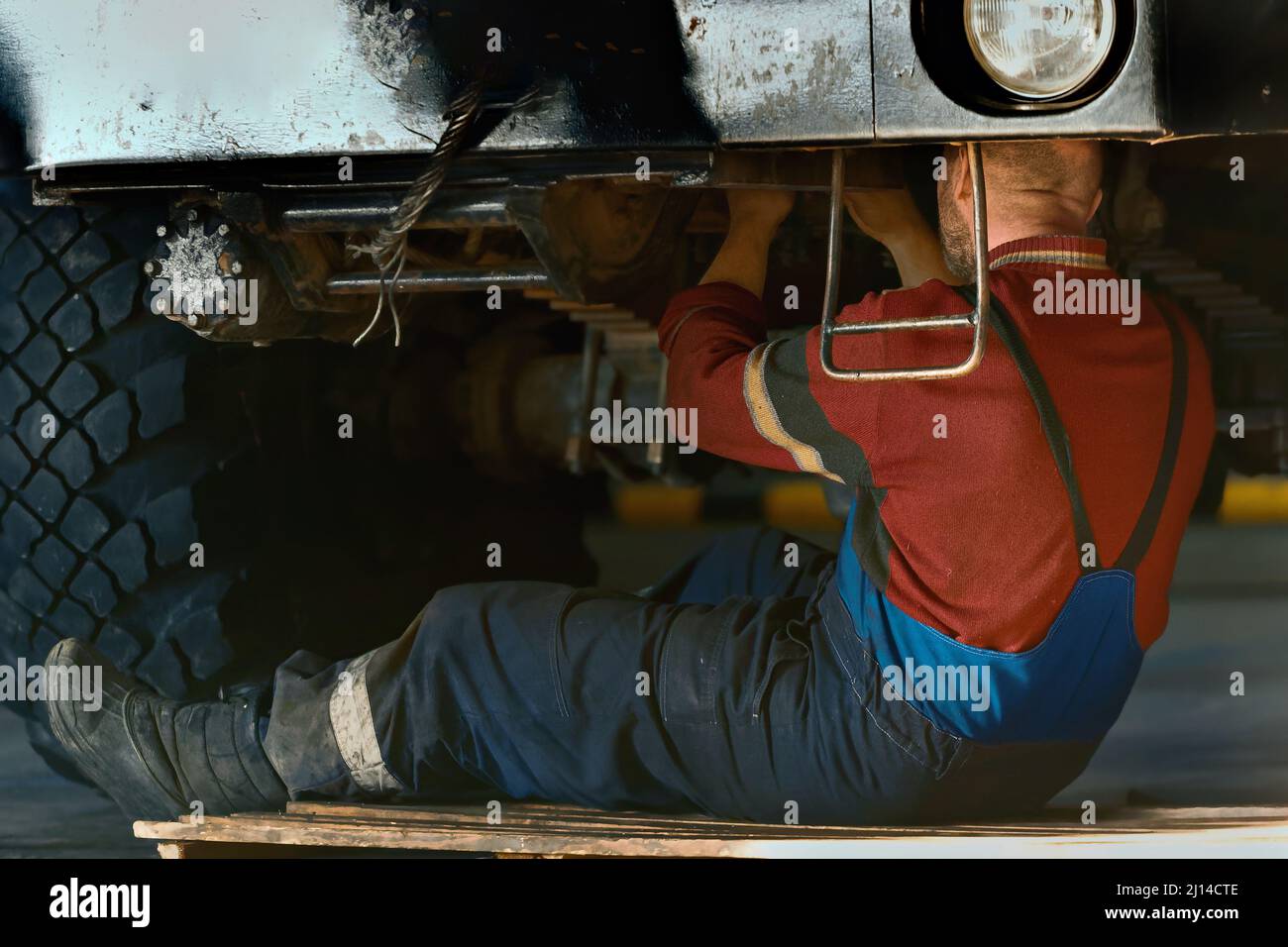 Il fabbro o meccanico dell'automobile siede sul pavimento sotto l'automobile e ripara l'apparecchiatura grande nel garage. Servizio auto. Sfondo. Foto Stock