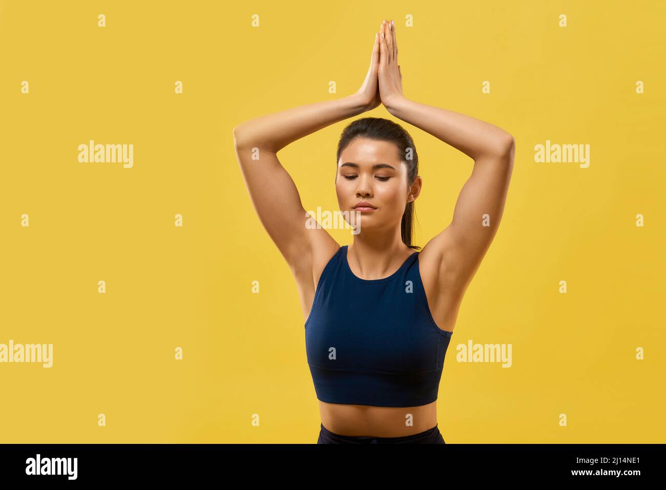 Primo piano della ragazza flessibile che tiene le mani di preghiera sopra la testa. Donna sportiva con coda di pony seduta, guardando giù, meditando, yoga fare. Isolato su yell Foto Stock