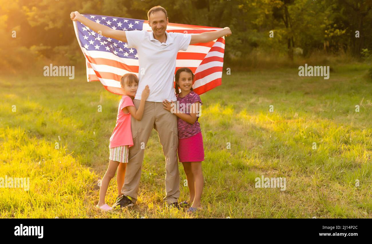 Felice famiglia seduta insieme nel loro cortile che tiene la bandiera americana dietro di loro. Coppia sorridente con i loro bambini che celebrano l'americano Foto Stock