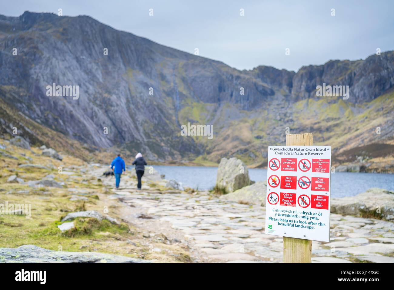 Vista posteriore della coppia che cammina sul lago nelle montagne gallesi del Parco Nazionale di Snowdonia; cartello in primo piano che informa il pubblico delle restrizioni della riserva naturale. Foto Stock