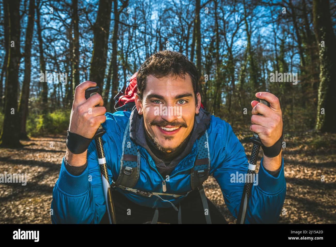 Giovane uomo sta facendo trekking nel bosco. Il bellissimo uomo ha una giacca blu, uno zaino rosso e bastoncini da trekking. Foto Stock