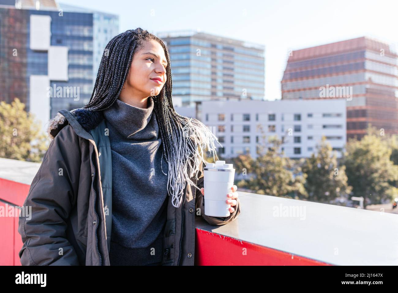 Attraente femmina ispanica in outerwear caldo con thermos di caffè caldo in piedi vicino al confine mentre osservano la città con edifici residenziali Foto Stock