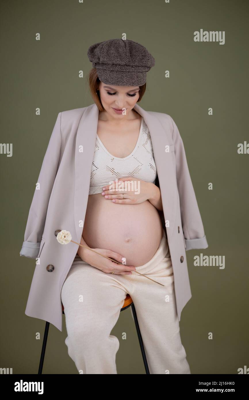 Giovane donna incinta sorridente con lunghi capelli scuri in cappello marrone, top bianco, cappotto grigio, pantaloni beige, craspedia di tenuta. Foto Stock