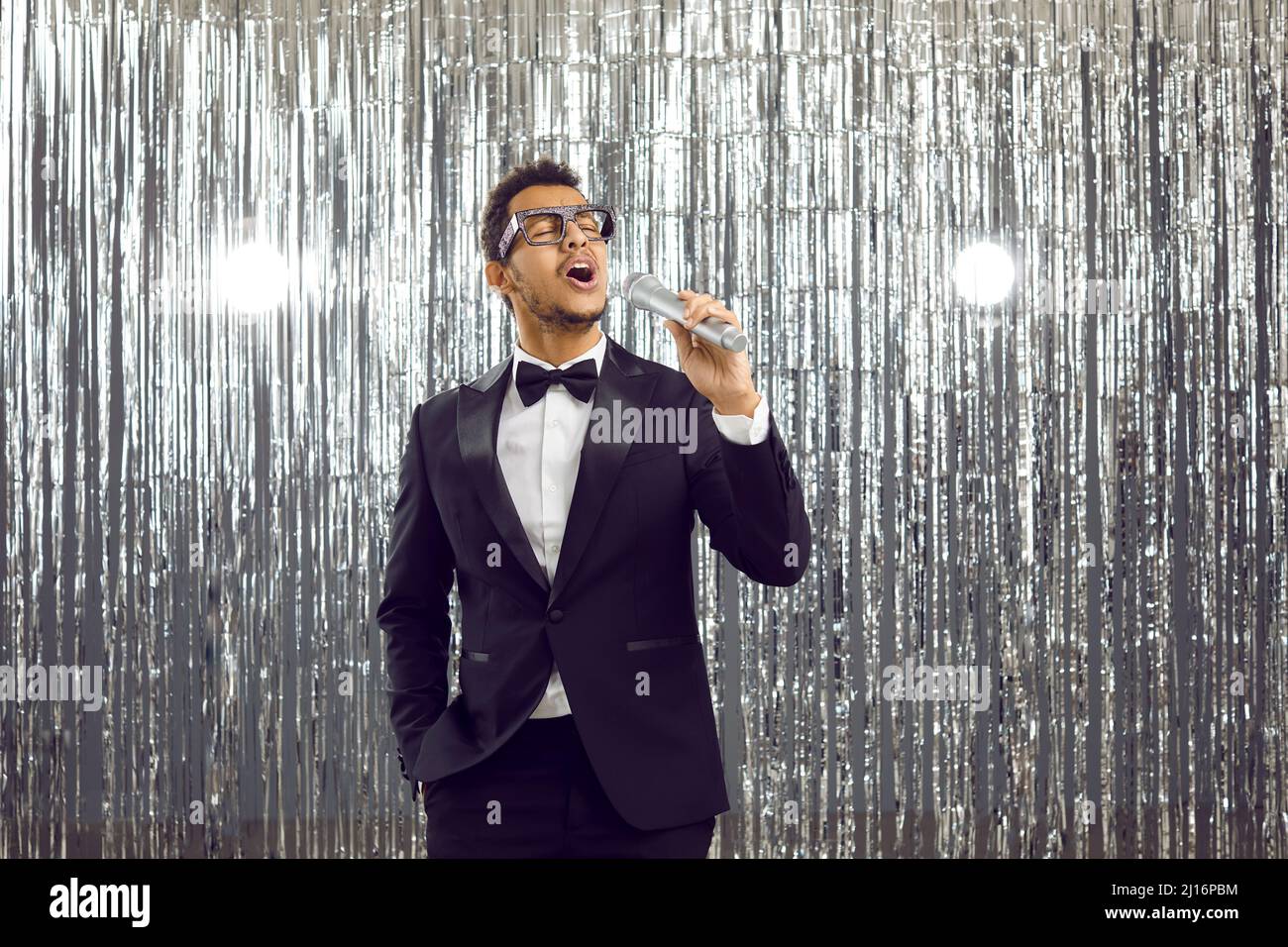 Giovane cantante nero in tuxedo vestito in piedi sul palco, tenendo microfono e cantando Foto Stock