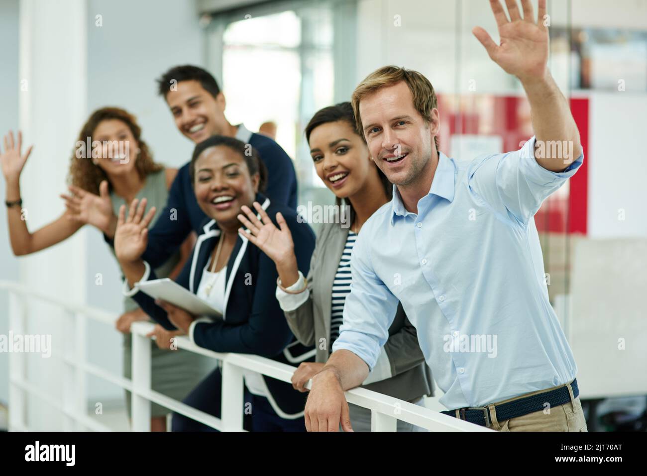 Ciao successo. Ritratto di un gruppo di colleghi sorridenti che ondeggiano mentre si appoggiano su una ringhiera in un ufficio. Foto Stock