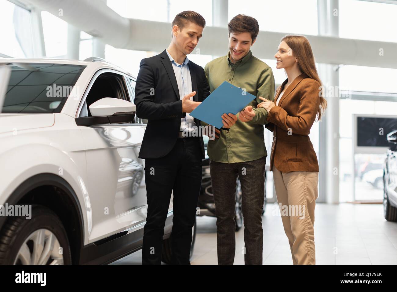 Un venditore di auto fiducioso che mostra i dettagli di acquisto alla giovane coppia caucasica presso il centro concessionaria Foto Stock