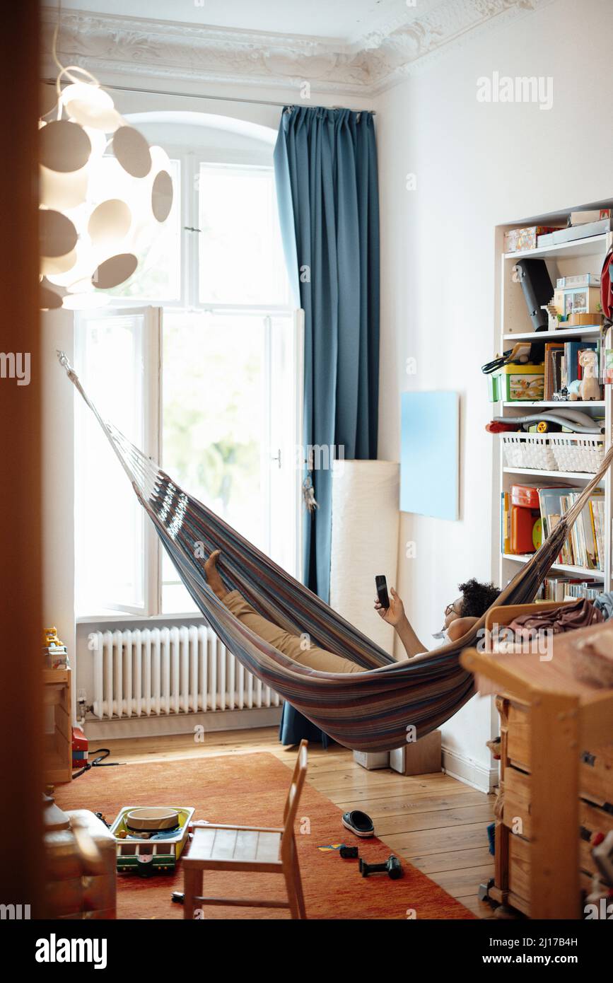 Uomo sdraiato in amaca utilizzando il telefono cellulare in soggiorno Foto Stock