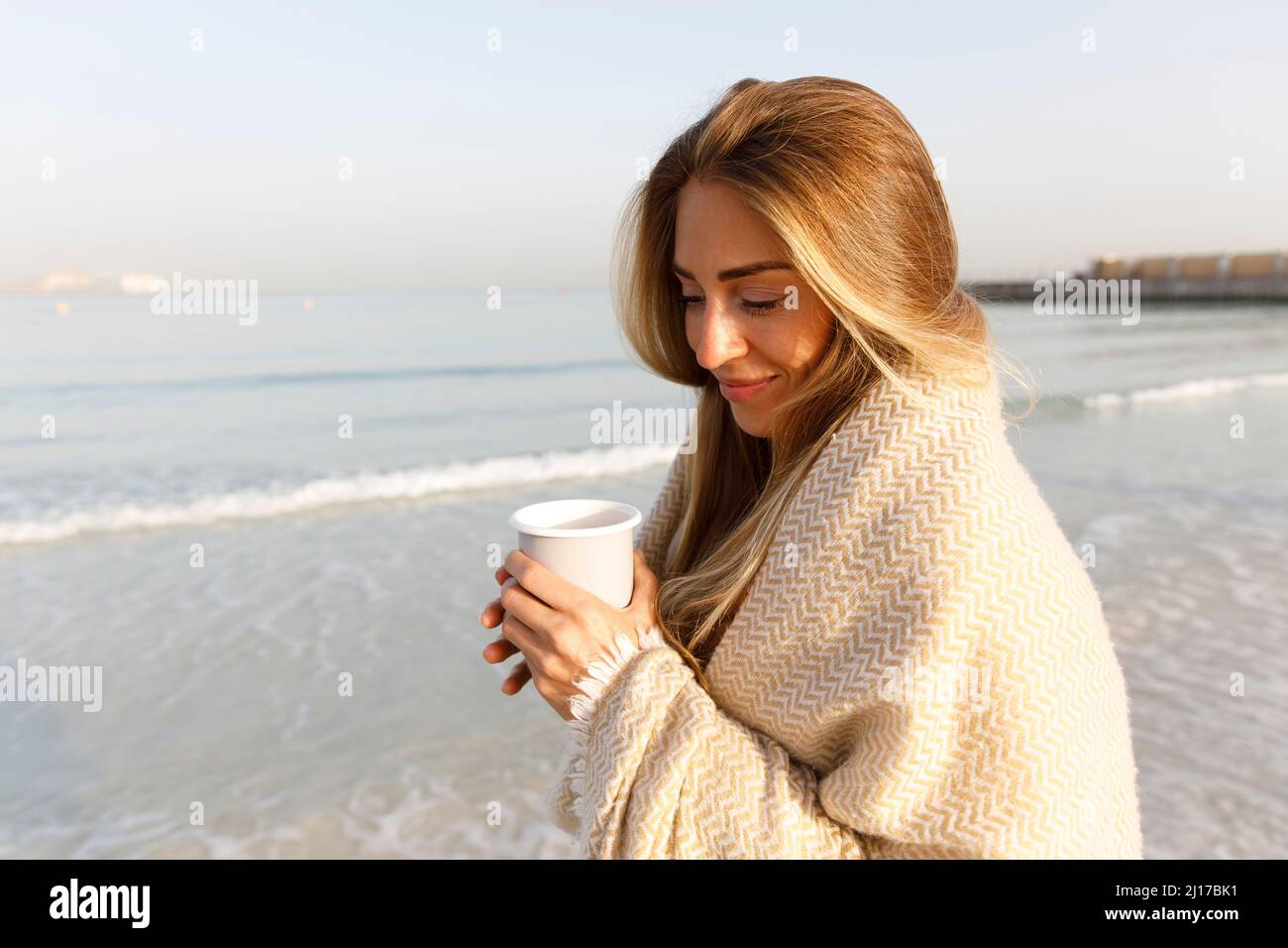 Donna bionda sorridente che tiene una tazza di tè in spiaggia Foto Stock